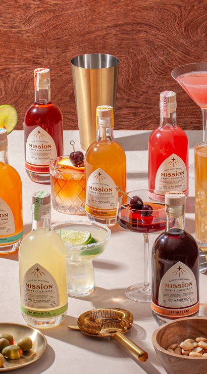 Vibrant spread of premixed craft cocktail bottles and mixed drinks on a tabletop, garnished with lime, cherries and pineapple, surrounded by barware, jigger, olives and nuts against a wood backdrop.