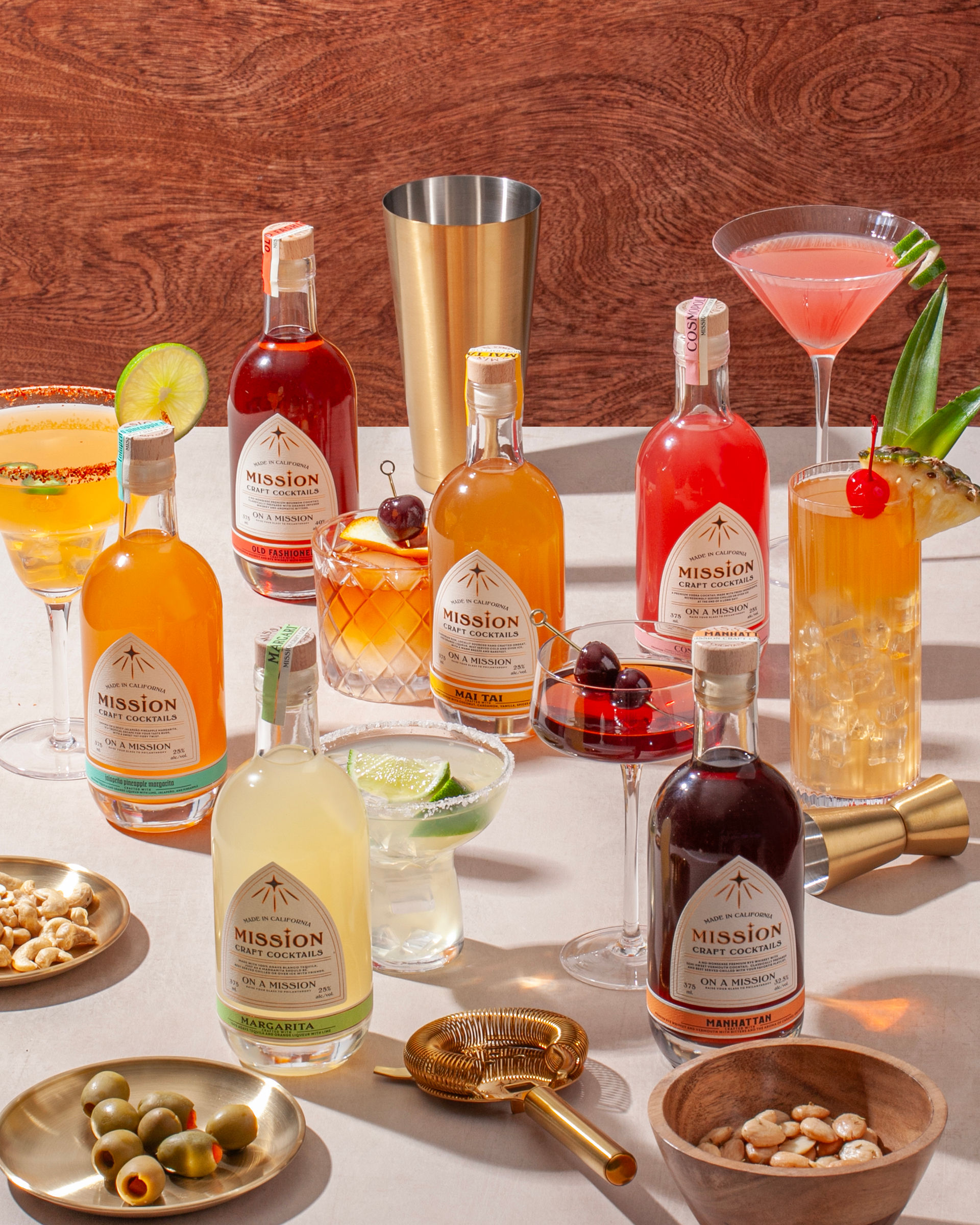 Vibrant spread of premixed craft cocktail bottles and mixed drinks on a tabletop, garnished with lime, cherries and pineapple, surrounded by barware, jigger, olives and nuts against a wood backdrop.