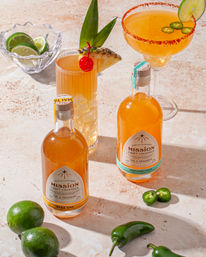 Sunlit stone tabletop with two bottles of amber craft cocktails, a tall tiki-style drink garnished with pineapple and cherry, and a jalapeño-rimmed margarita, surrounded by limes and sliced jalapeños.