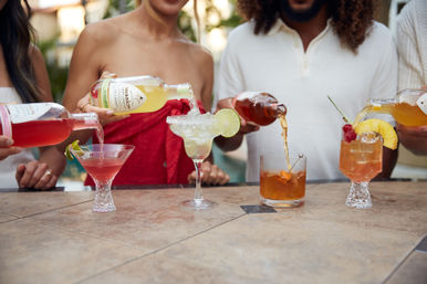 Four colorful cocktails being poured on an outdoor patio bar—pink martini with a lime twist, salted-rim margarita garnished with a lime wheel, amber old-fashioned with an orange peel, and a pineapple-cherry tropical glass.