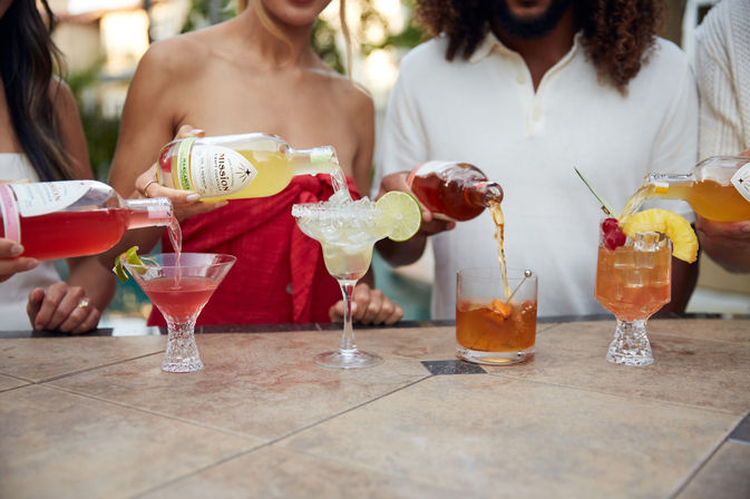 Four colorful cocktails being poured on an outdoor patio bar—pink martini with a lime twist, salted-rim margarita garnished with a lime wheel, amber old-fashioned with an orange peel, and a pineapple-cherry tropical glass.