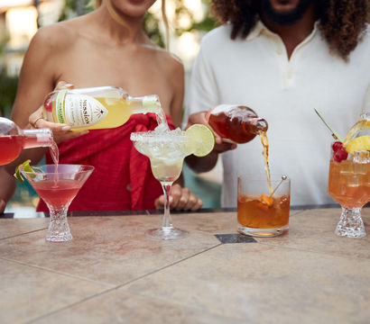 Four colorful cocktails being poured on an outdoor patio bar—pink martini with a lime twist, salted-rim margarita garnished with a lime wheel, amber old-fashioned with an orange peel, and a pineapple-cherry tropical glass.