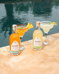 Sunny poolside scene with two bottled cocktails and matching margarita glasses — pineapple-chili rim and salted lime wedge on a stone deck.