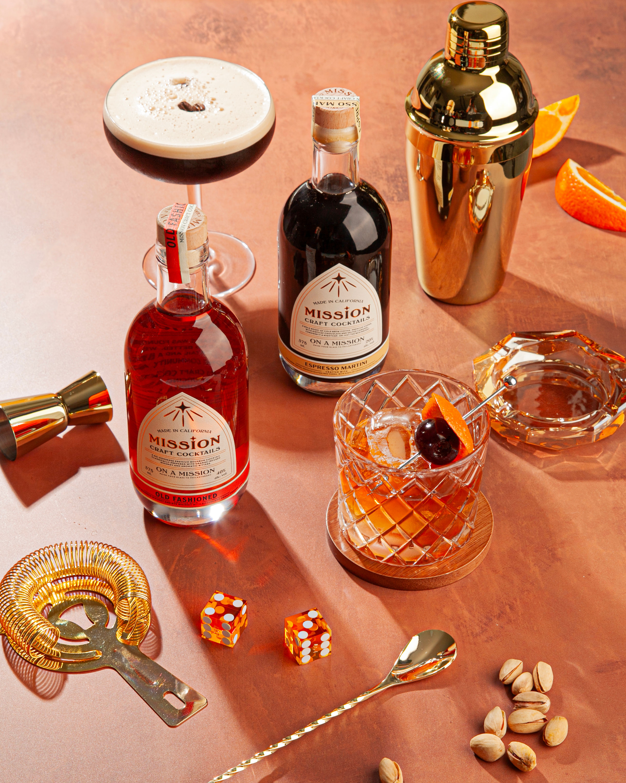 Copper tabletop home bar scene with two bottles of craft cocktails, an espresso martini coupe, a crystal old-fashioned with cherry and orange peel, gold shaker and bar tools, and a handful of pistachios.