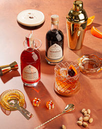 Copper tabletop home bar scene with two bottles of craft cocktails, an espresso martini coupe, a crystal old-fashioned with cherry and orange peel, gold shaker and bar tools, and a handful of pistachios.