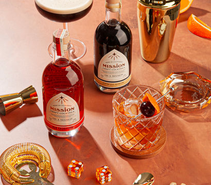 Copper tabletop home bar scene with two bottles of craft cocktails, an espresso martini coupe, a crystal old-fashioned with cherry and orange peel, gold shaker and bar tools, and a handful of pistachios.