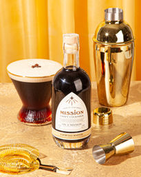 Bottle of espresso martini cocktail with a crema‑topped martini glass, gold cocktail shaker, jigger and spring strainer arranged on a shimmering gold tabletop with a golden curtain backdrop.
