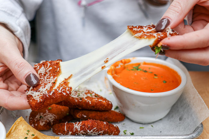 Close-up of hands pulling a crispy breaded mozzarella stick apart, gooey cheese stretching toward a ramekin of marinara sauce on a parchment-lined tray.