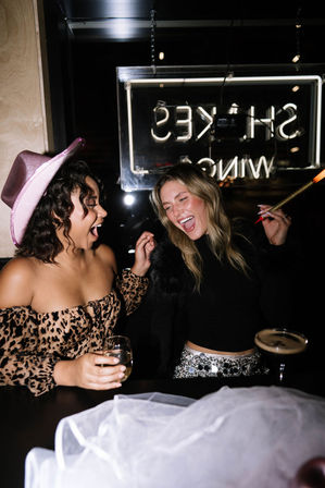 Two friends laughing at a city bar, one wearing a pink cowboy hat and leopard-print top holding a wine glass, the other waving a sparkler by a cocktail under a neon sign.