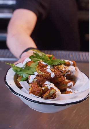 Crispy battered vegetable bites drizzled with creamy ranch and paprika, garnished with fresh parsley in a white enamel bowl being handed across a restaurant counter