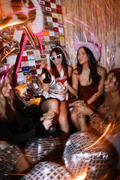 Four friends at a lively indoor bachelorette/girls' night party — one in heart-shaped sunglasses popping champagne, another in a pink cowboy hat, all holding cocktails amid disco balls, metallic tinsel backdrop and a colorful cassette-tape wall.