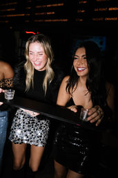 Two women laughing on a night out at a nightclub, holding shot glasses on a shared tray — one wearing a silver sequin mini skirt and black top, the other in a black sequin dress.