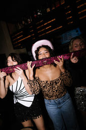 Three friends at a dim bar sharing a pink shotski and laughing — center woman in a pink cowboy hat and leopard-print top, left in a black-and-white fringe dress, right in a sequined skirt on a lively night out.