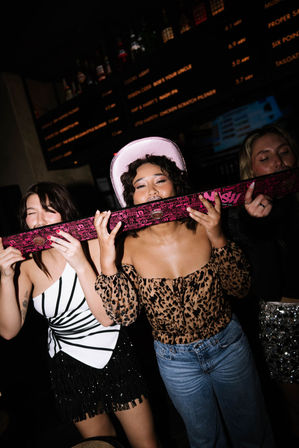 Three friends at a dim bar sharing a pink shotski and laughing — center woman in a pink cowboy hat and leopard-print top, left in a black-and-white fringe dress, right in a sequined skirt on a lively night out.