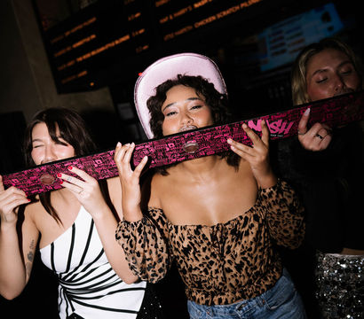 Three friends at a dim bar sharing a pink shotski and laughing — center woman in a pink cowboy hat and leopard-print top, left in a black-and-white fringe dress, right in a sequined skirt on a lively night out.