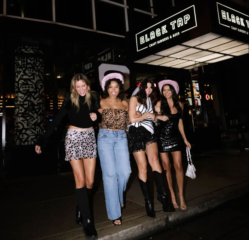 Four friends arm-in-arm on a downtown night out, laughing in party outfits—sequins, boots, jeans—with two wearing pink cowgirl hats under bright restaurant lights