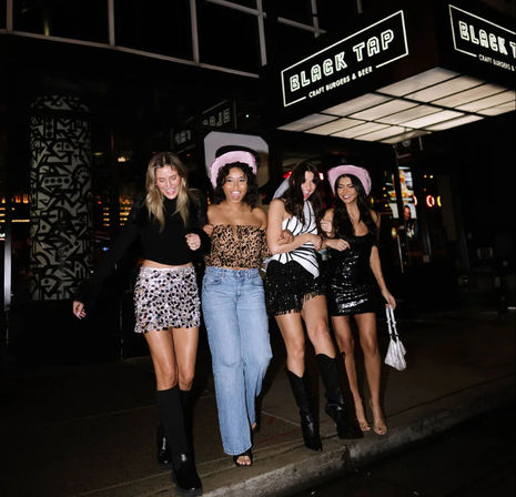 Four friends arm-in-arm on a downtown night out, laughing in party outfits—sequins, boots, jeans—with two wearing pink cowgirl hats under bright restaurant lights