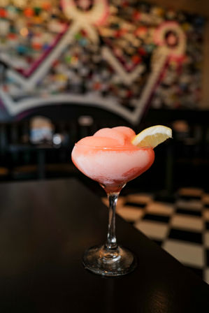 Pink frozen cocktail in a stemmed coupe glass with a lemon wedge garnish on a black table, blurred colorful mural and black-and-white tiled bar floor in the background.