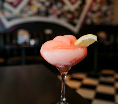 Pink frozen cocktail in a stemmed coupe glass with a lemon wedge garnish on a black table, blurred colorful mural and black-and-white tiled bar floor in the background.