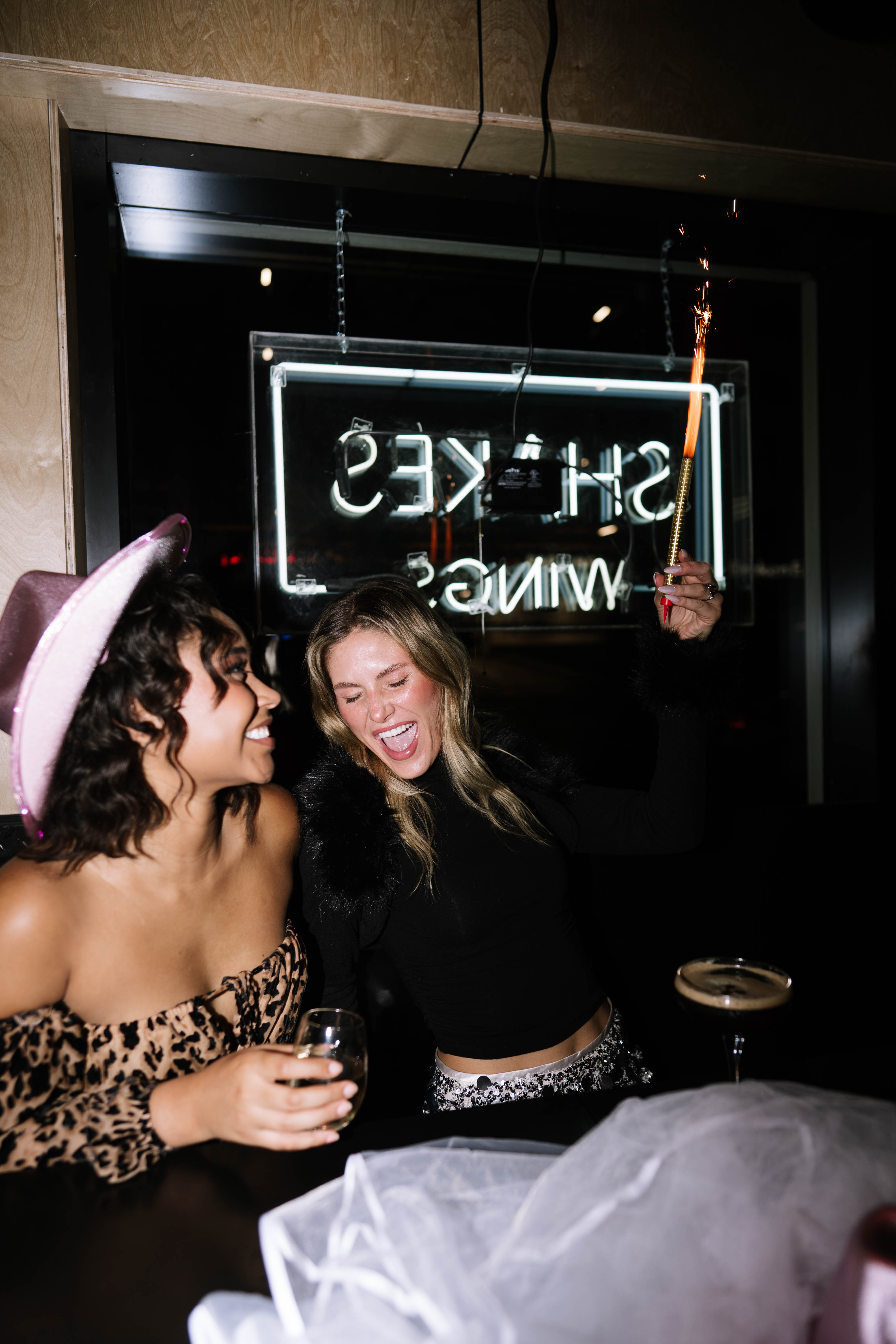 Two friends laughing at a bar at night — one wearing a pink cowboy hat and leopard off-shoulder top holding a wine glass, the other in a black top waving a lit sparkler with neon sign and cocktails behind them.