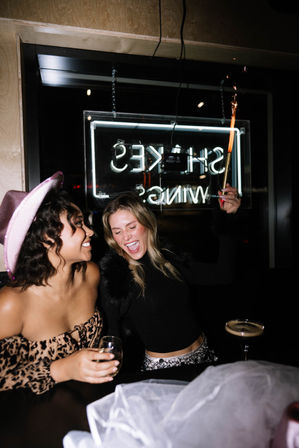 Two friends laughing at a bar at night — one wearing a pink cowboy hat and leopard off-shoulder top holding a wine glass, the other in a black top waving a lit sparkler with neon sign and cocktails behind them.
