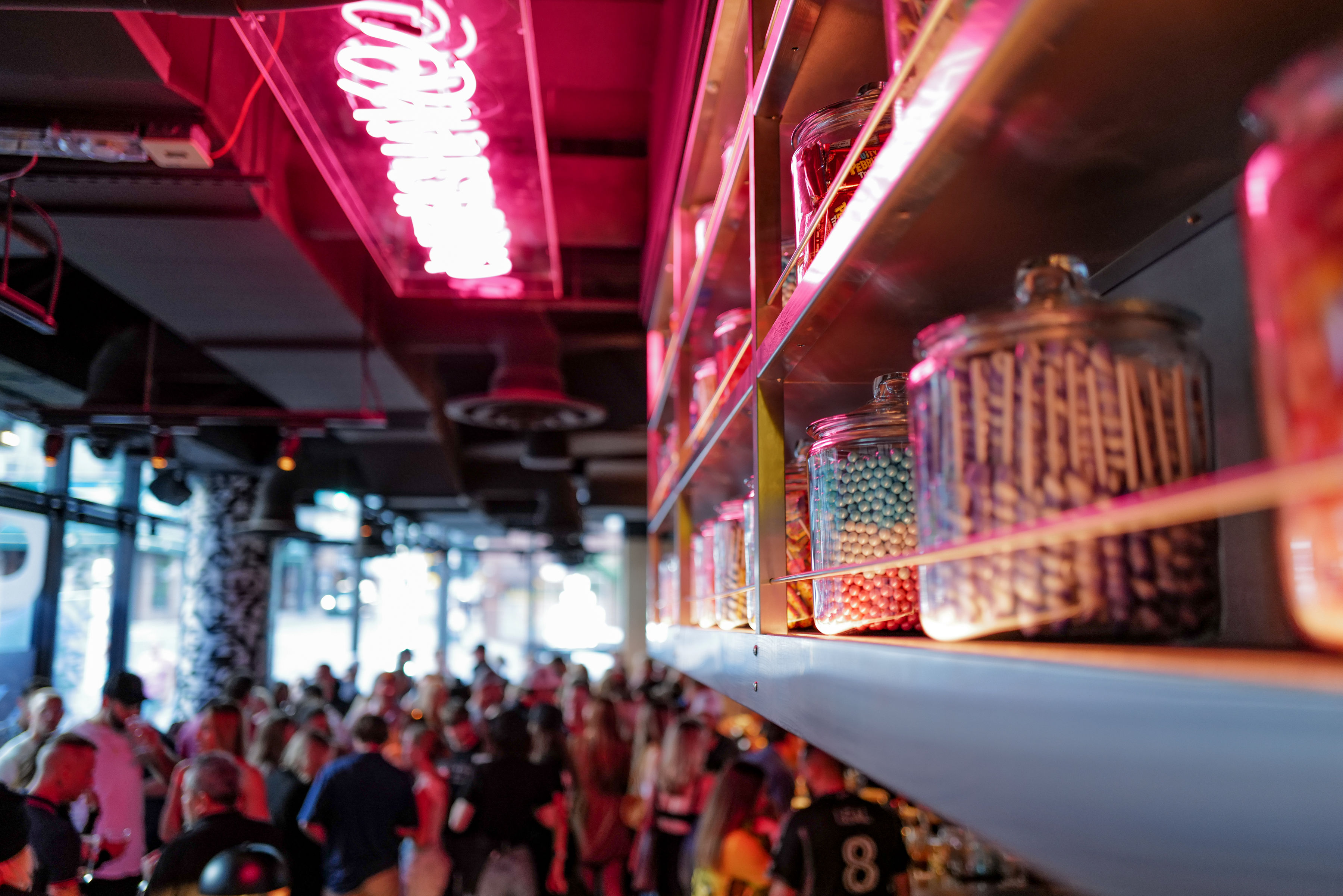 Neon-lit indoor venue with a pink neon sign, glass jars of colorful candy on a shelf in sharp focus and a blurred crowd mingling below