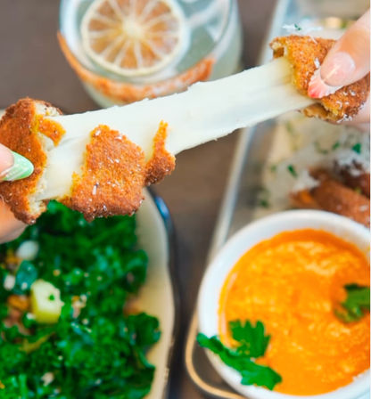 Close-up of hands pulling apart a crispy breaded mozzarella stick with a gooey cheese stretch, plated with orange dipping sauce, a leafy kale salad and a lemon-slice drink.