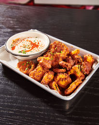 Crispy spiced fried cauliflower bites piled on a metal tray with creamy paprika-dusted dipping sauce and chopped parsley on a dark wood table, savory appetizer