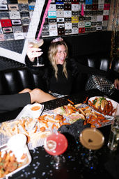 Woman wearing a party tiara toasting with a cocktail in a retro-themed nightclub booth with cassette-tape wall art, plates of fries and shared bar snacks on the table.