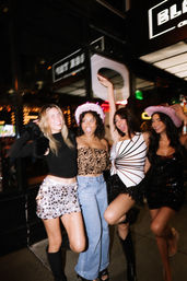 Four friends partying on a downtown sidewalk at night, wearing sparkly outfits and pink cowboy hats outside a lit-up bar — city nightlife scene
