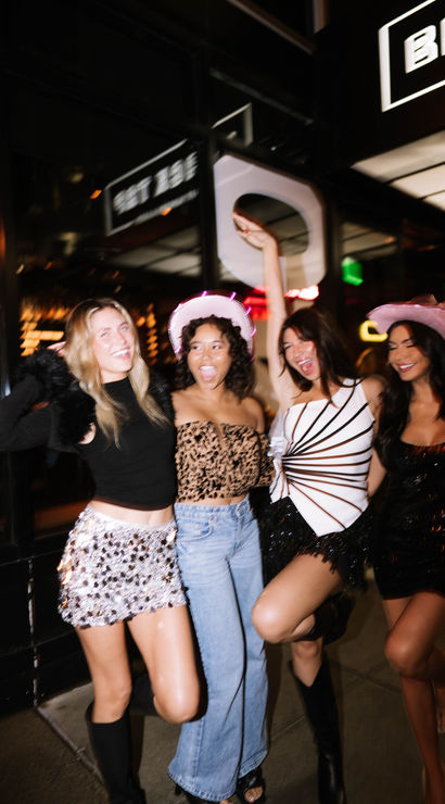 Four friends partying on a downtown sidewalk at night, wearing sparkly outfits and pink cowboy hats outside a lit-up bar — city nightlife scene