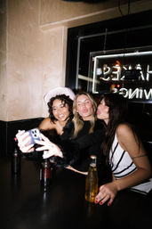 Three friends taking a selfie during a night out at a trendy urban bar booth, neon sign glowing in the window and beer bottles on the table; one wears a pink cowboy hat.