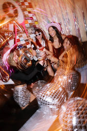 Four friends toasting cocktails and champagne at a glittery indoor party with disco balls, tinsel backdrop, and colorful light streaks.
