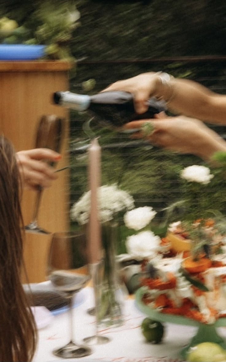 Hands pouring sparkling wine into a glass at an al fresco garden dinner table with candles, white flowers and a colorful cake stand — motion-blurred, summery celebration vibe.