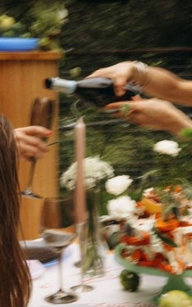Hands pouring sparkling wine into a glass at an al fresco garden dinner table with candles, white flowers and a colorful cake stand — motion-blurred, summery celebration vibe.