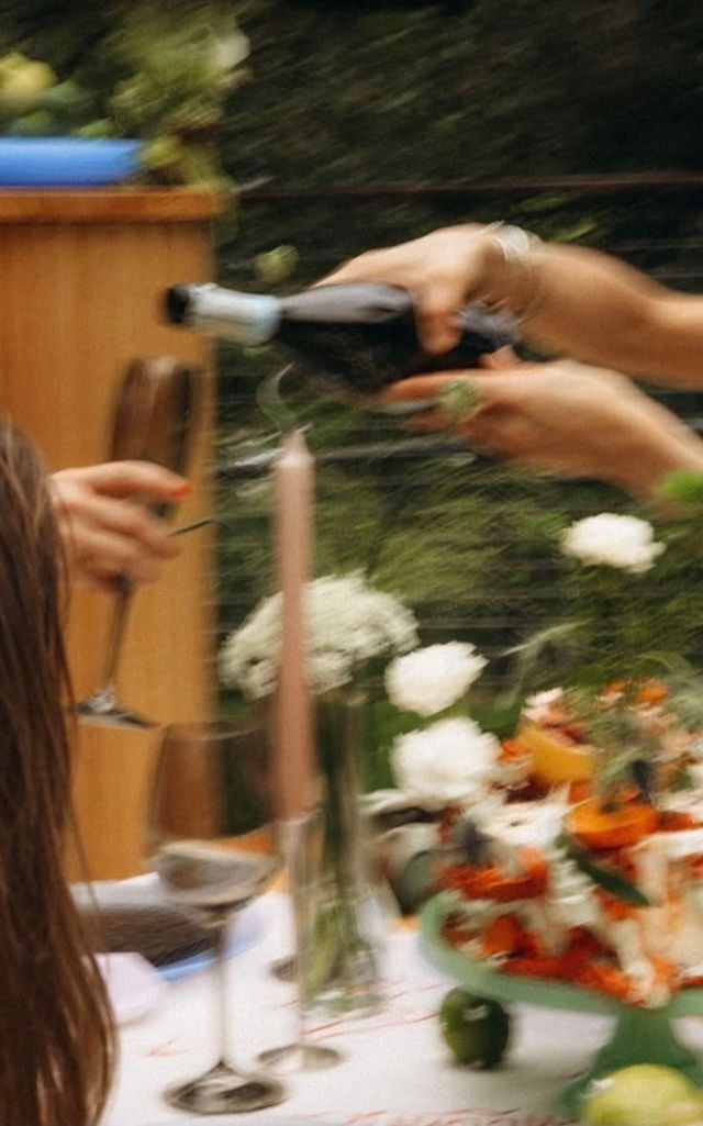 Hands pouring sparkling wine into a glass at an al fresco garden dinner table with candles, white flowers and a colorful cake stand — motion-blurred, summery celebration vibe.