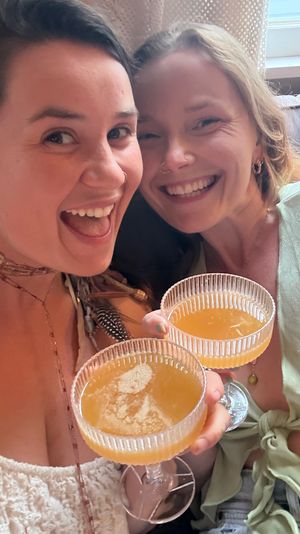 Close-up selfie of two friends on a couch near a window, smiling and toasting with coupe glasses of orange cocktails — cheers!