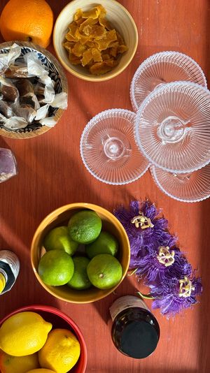Overhead flat-lay of a wooden tray with bowls of limes and lemons, glass coasters, wrapped caramels, candied orange peel, purple passionflowers and an amethyst — colorful kitchen still life.