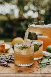 Pouring refreshing lavender-mint iced tea from a swing-top glass bottle into textured tumblers on a wooden table in a sunlit garden.