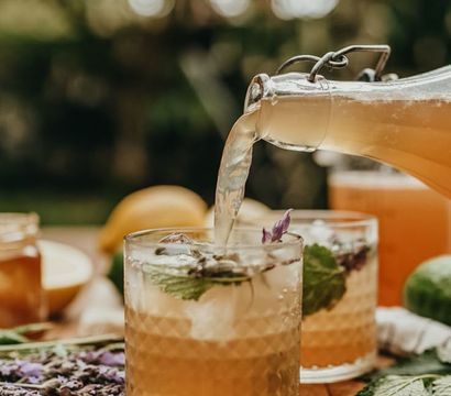 Pouring refreshing lavender-mint iced tea from a swing-top glass bottle into textured tumblers on a wooden table in a sunlit garden.