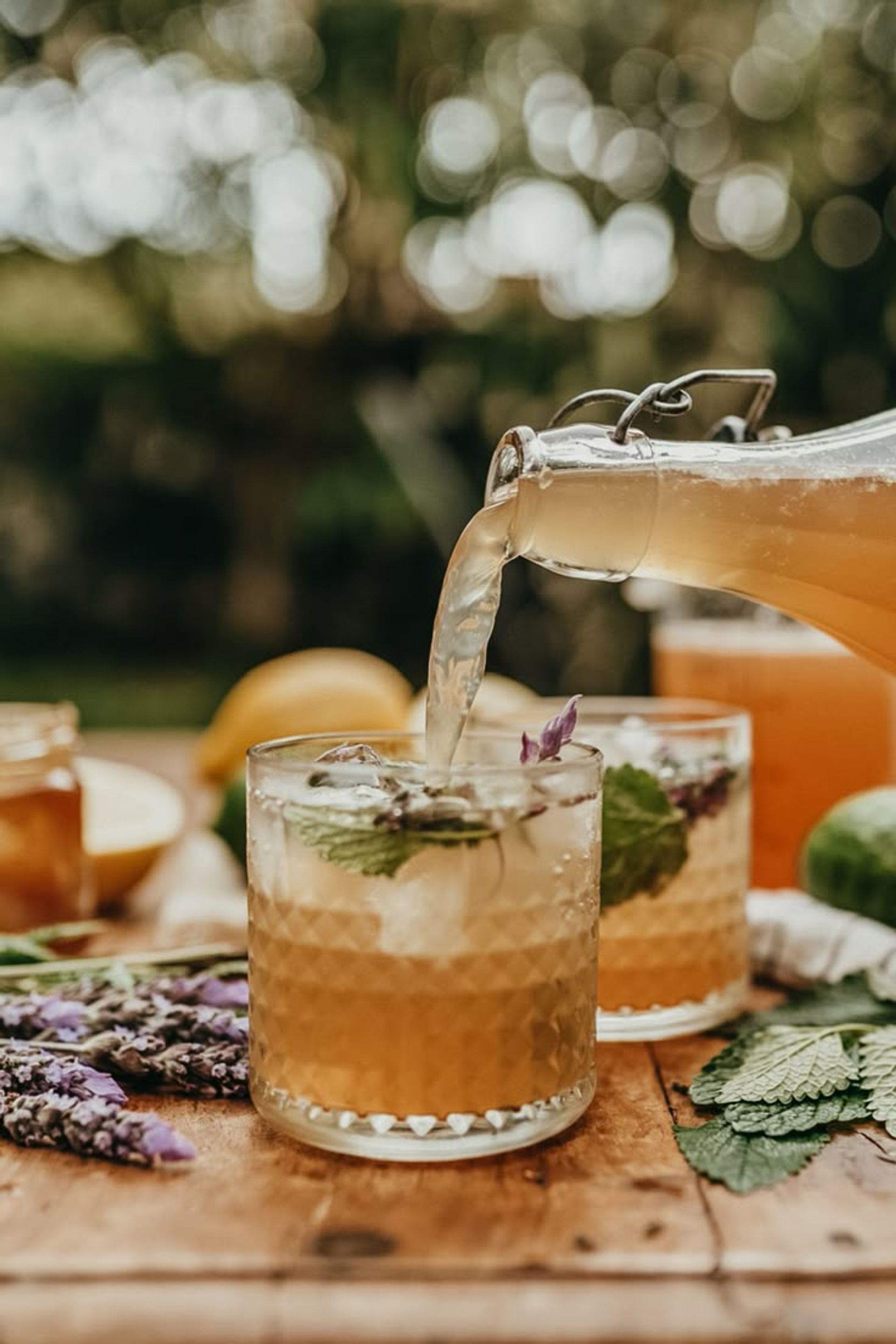 Pouring refreshing lavender-mint iced tea from a swing-top glass bottle into textured tumblers on a wooden table in a sunlit garden.