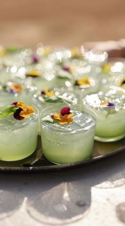 Sunlit tray of lime-green iced cocktails in clear cups, garnished with edible pansy flowers and green leaves at an outdoor reception