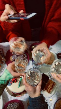 Overhead view of a small celebration: five hands clinking crystal glasses above a table of cake, cookies and sweets, one person photographing the moment on a smartphone.