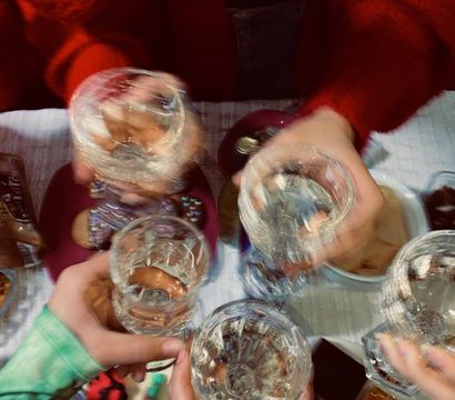 Overhead view of a small celebration: five hands clinking crystal glasses above a table of cake, cookies and sweets, one person photographing the moment on a smartphone.