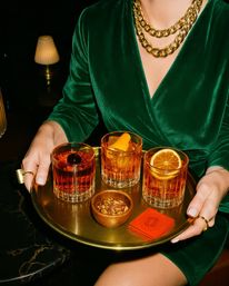 Person in a green velvet dress and chunky gold necklace holds a brass tray with three amber cocktails in old‑fashioned glasses (cherry, orange peel, orange wheel), a small bowl of nuts and red napkins in a dim lounge setting.