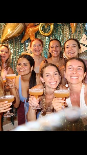 Smiling group of women raising coupe cocktails in front of a teal sequin mermaid-style backdrop with gold starfish balloons — festive girls' night or bachelorette celebration indoors.