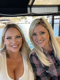 Two smiling blonde women taking a selfie on a covered boat with a waterfront marina, docked yachts and bright blue harbor water in the background