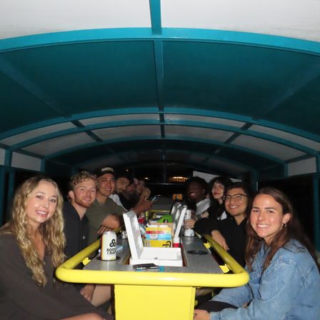 Smiling group of young adults seated around a yellow bar inside a covered party bike at night, enjoying drinks and takeout containers