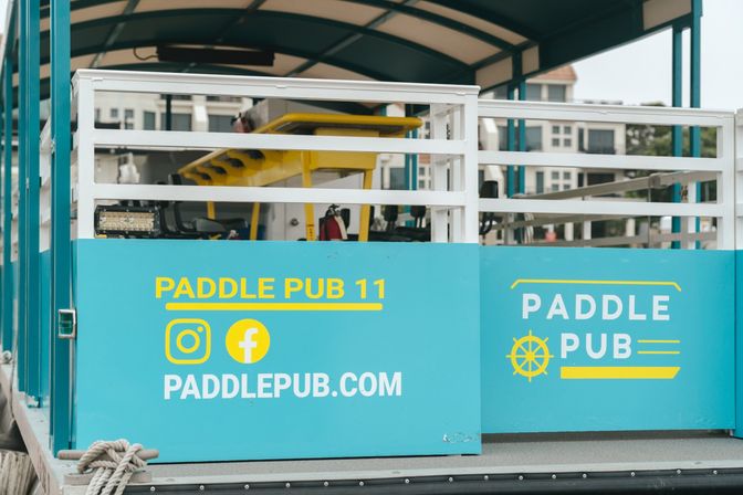Teal pedal-powered party boat docked at a marina with white railings, yellow seating and social media icons on the side panel.