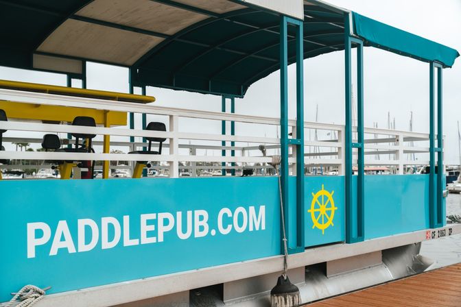 Teal covered paddle barge docked at a marina with bar-style seats, a yellow ship wheel graphic, and boats in the background